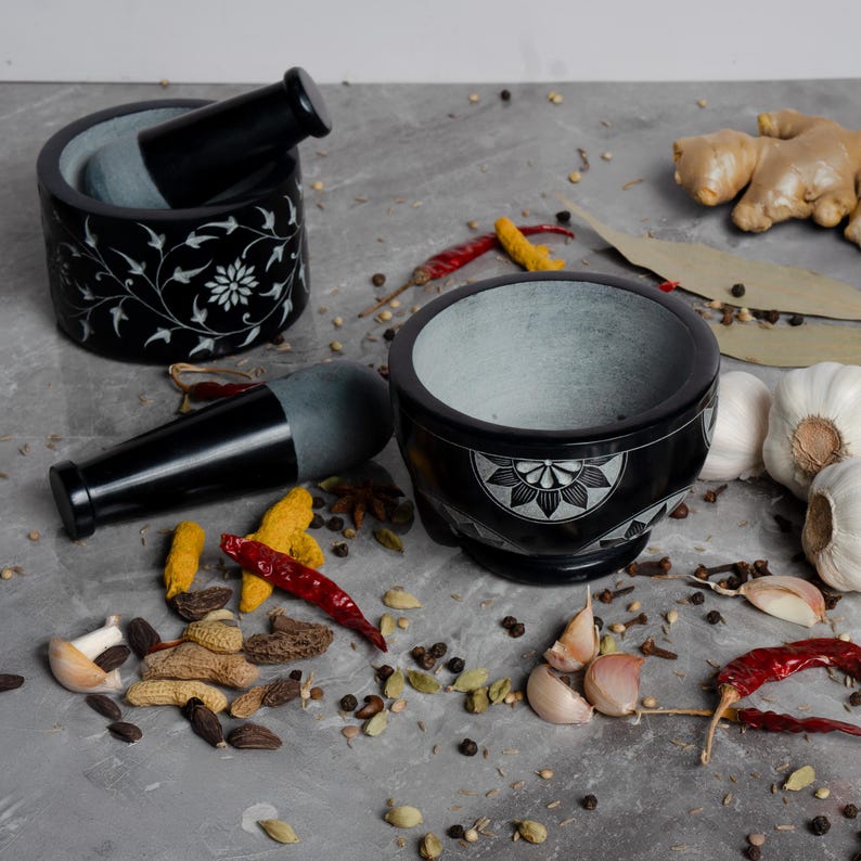 May include: Two black stone mortar and pestle sets with white floral designs. One mortar is cylindrical, the other is bowl-shaped. Spices, garlic, and ginger are scattered around the sets on a gray surface.