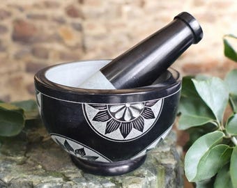 May include: A black mortar and pestle set with a floral design carved into the exterior. The interior of the bowl is white. The pestle is black with a white grinding end. The set is made of stone and sits on a stone surface.
