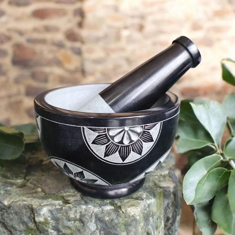 May include: A black mortar and pestle set with a floral design carved into the exterior. The interior of the bowl is white. The pestle is black with a white grinding end. The set is made of stone and sits on a stone surface.