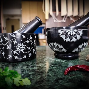 May include: Two black mortar and pestle sets with white floral designs. One is cylindrical with a floral and vine pattern, the other is bowl-shaped with a pedestal base. A red chili pepper and cilantro are in the foreground.