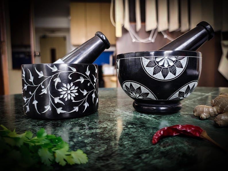 May include: Two black mortar and pestle sets with white floral designs. One is cylindrical with a floral and vine pattern, the other is bowl-shaped with a pedestal base. A red chili pepper and cilantro are in the foreground.