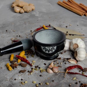 May include: A black mortar and pestle set with a floral design, surrounded by various spices like garlic, ginger, cinnamon sticks, and red chilies. The mortar is made of stone and has a gray interior.
