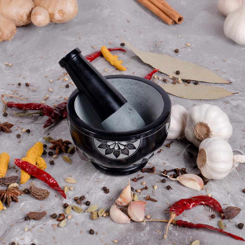 May include: A black mortar and pestle set with a floral design on the exterior. The mortar is filled with a gray interior. Surrounding the mortar are various spices, including red chilies, garlic cloves, and peppercorns.
