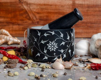 May include: A black mortar and pestle with a floral design, alongside various spices and garlic. The mortar is cylindrical, with a matching pestle resting inside. Red chili peppers, ginger, and peppercorns are scattered around the mortar.