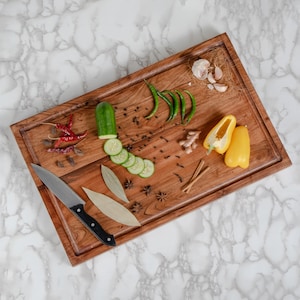 May include: A rectangular wooden cutting board with a knife, sliced cucumber, yellow bell peppers, green chilies, and spices. The board has a carved border and is set against a marble background.