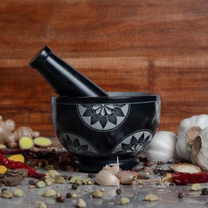 May include: A black mortar and pestle set with a floral design, resting on a surface with various spices and garlic. The pestle is angled inside the bowl. The background is a wooden surface.