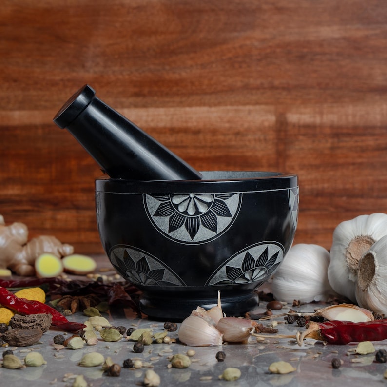 May include: A black mortar and pestle set with a floral design, resting on a surface with various spices and garlic. The pestle is angled inside the bowl. The background is a wooden surface.