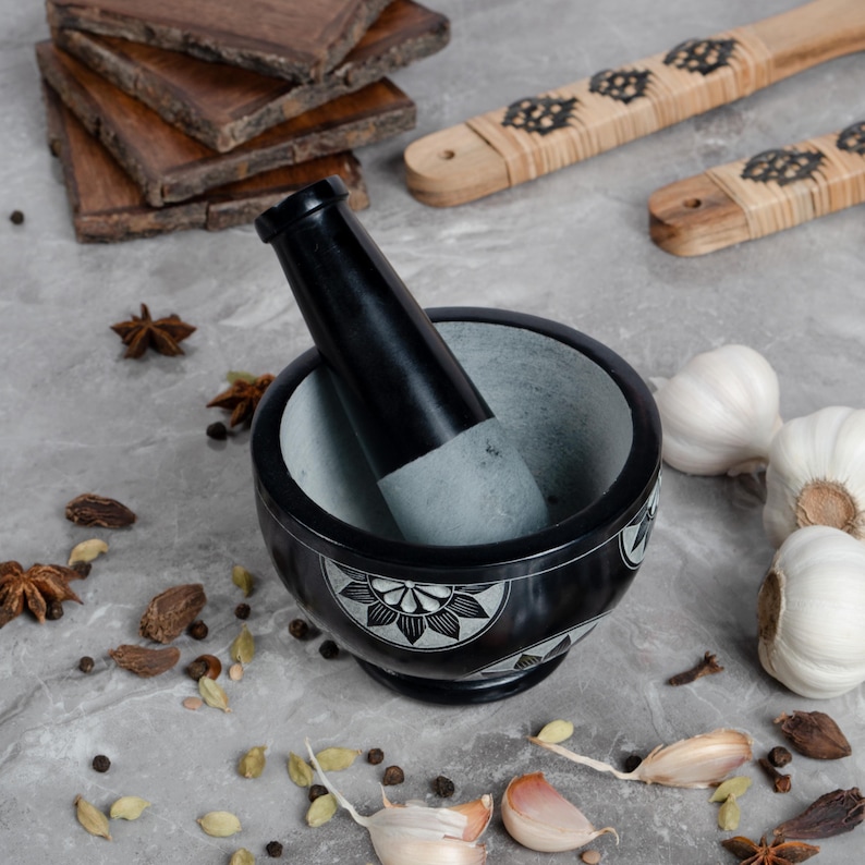 May include: A black and gray mortar and pestle set with a floral design. The mortar is made of stone and the pestle is black. The image also shows wooden serving boards, garlic, and spices.