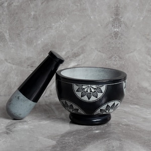 May include: A black mortar and pestle set. The mortar is round with a pedestal base and floral design. The pestle is black with a gray tip. The set is made of stone and is suitable for grinding herbs and spices.
