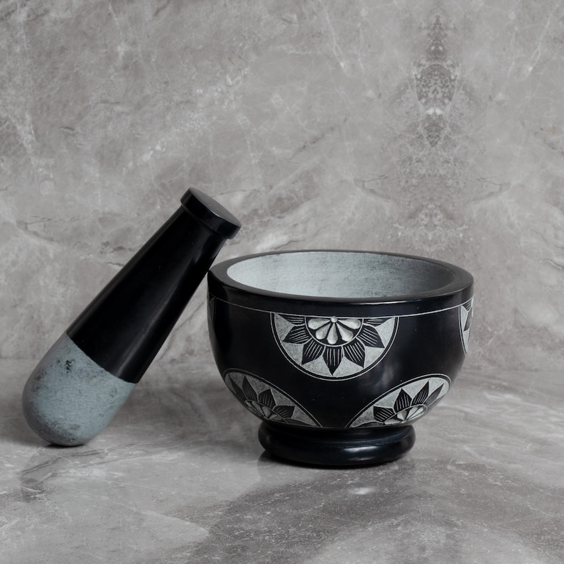May include: A black mortar and pestle set. The mortar is round with a pedestal base and floral design. The pestle is black with a gray tip. The set is made of stone and is suitable for grinding herbs and spices.