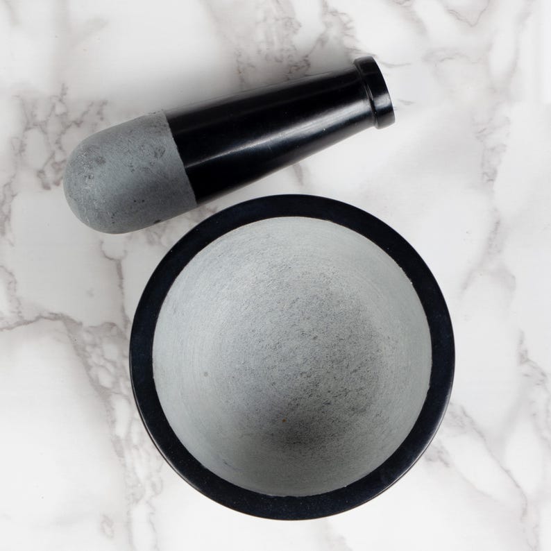 May include: A black and gray mortar and pestle set. The mortar is a gray bowl with a black rim. The pestle is black with a gray grinding end. The set is on a white and gray marble surface.