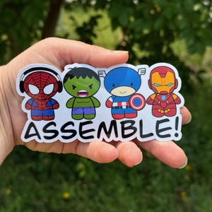 May include: A sticker featuring four cartoon superheroes: Spider-Man, Hulk, Captain America, and Iron Man. The word "ASSEMBLE!" is printed below the characters. The sticker is held in a hand against a blurred green background.