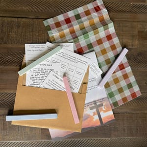 May include: A collection of printed cards, a brown envelope, and four slim rectangular pens in light green, pink, light blue, and lavender. A sheet of paper features a cross silhouette at sunset and the text begin again.