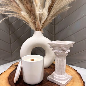 Può includere: Una candela in ceramica bianca con uno stoppino di legno si trova su una fetta di legno accanto a un vaso in ceramica bianca con un foro al centro e una colonna in gesso bianca con dettagli decorativi.