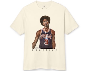 May include: A cream-colored short-sleeve t-shirt featuring a graphic illustration of a basketball player wearing a dark blue jersey with the text PHLA and the number 3. Below the illustration, the word PRACTICE is printed in black capital letters.