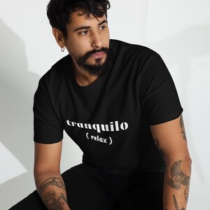 May include: Black t-shirt with the word "tranquilo" and the word "(relax)" printed in white. The t-shirt is made of a soft material and has a classic crew neck design. The shirt is worn by a person.