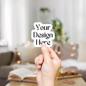 Puede incluir: Una pegatina blanca con el texto "Your Design Here" en ella. La pegatina está siendo sostenida por una mano contra un fondo borroso de una sala de estar.