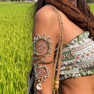 May include: A silver arm cuff with a spiral design and a seashell charm. The cuff is adorned with small green beads and wire detailing. The arm cuff is worn with a green and white floral print top and a brown scarf.