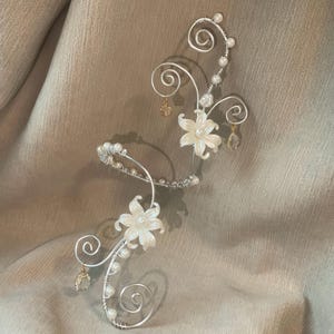 May include: A silver-toned, floral-inspired hair accessory. The piece features delicate wirework, adorned with small pearls and two white flower accents. Small, dangling crystal and gold-toned charms add a touch of sparkle. The design includes elegant swirls.