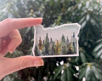 Puede incluir: Una pegatina de acuarela del estado de Oregón con una escena de bosque. La pegatina es blanca con un cielo azul claro y árboles verdes.
