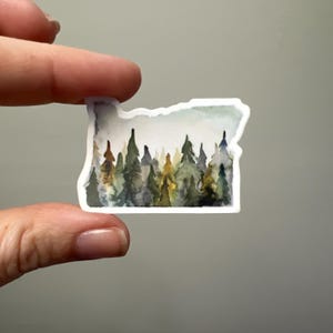 May include: A watercolor sticker of the state of Oregon with a forest scene. The sticker is white with a light blue sky and green, brown, and yellow trees.