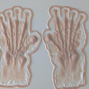 May include: A pair of prosthetic hands, featuring a detailed, textured design. The hands are a fleshy pink colour with a white border, and are likely for costume or theatrical use.