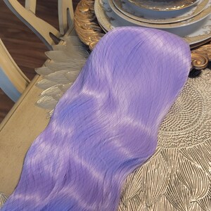 Puede incluir: Una peluca sint&eacute;tica larga y ondulada de un vibrante color morado lavanda. Las fibras capilares son lisas y tienen un ligero brillo, con una fina redecilla que cubre la parte superior de la peluca para mantener su forma y textura.