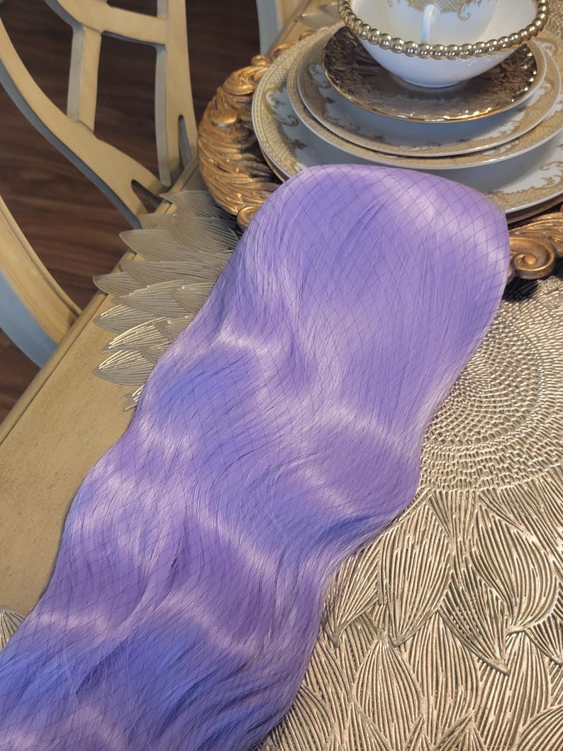 Puede incluir: Una peluca sint&eacute;tica larga y ondulada de un vibrante color morado lavanda. Las fibras capilares son lisas y tienen un ligero brillo, con una fina redecilla que cubre la parte superior de la peluca para mantener su forma y textura.
