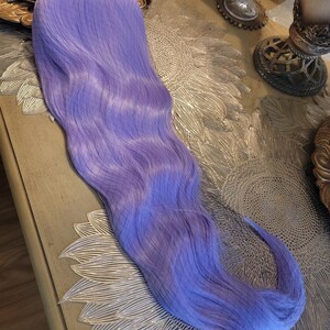 Puede incluir: Una peluca sint&eacute;tica larga y ondulada de un vibrante color morado lavanda. Las fibras capilares son lisas y est&aacute;n peinadas con ondas suaves y fluidas que se extienden desde la parte superior hasta las puntas, creando un aspecto uniforme.