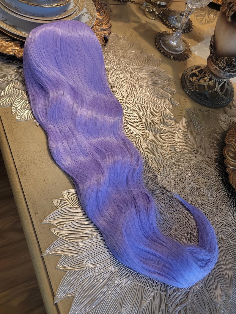 Puede incluir: Una peluca sint&eacute;tica larga y ondulada de un vibrante color morado lavanda. Las fibras capilares son lisas y est&aacute;n peinadas con ondas suaves y fluidas que se extienden desde la parte superior hasta las puntas, creando un aspecto uniforme.