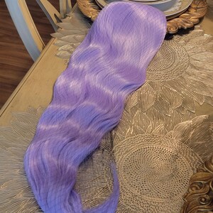 Puede incluir: Una peluca sint&eacute;tica larga y ondulada de un vibrante color morado lavanda. La pieza cuenta con una construcci&oacute;n de gorro con frente de encaje transparente y una malla fina visible en la base para una apariencia de l&iacute;nea de cabello natural.