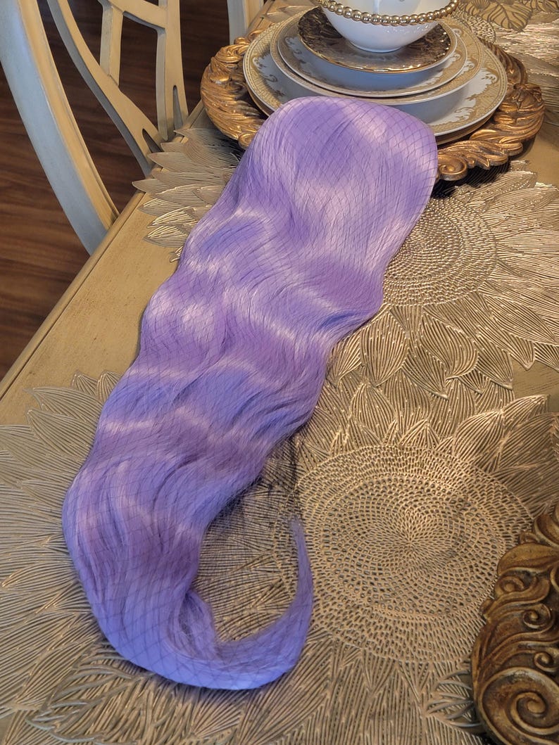 Puede incluir: Una peluca sint&eacute;tica larga y ondulada de un vibrante color morado lavanda. La pieza cuenta con una construcci&oacute;n de gorro con frente de encaje transparente y una malla fina visible en la base para una apariencia de l&iacute;nea de cabello natural.