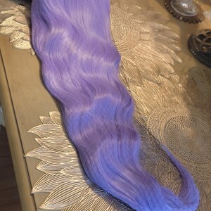 Puede incluir: Una peluca larga y ondulada de un vibrante color morado lavanda. Las fibras capilares son lisas y est&aacute;n peinadas con ondas suaves y fluidas que se extienden desde la coronilla hasta las puntas, creando una textura uniforme en toda la longitud.