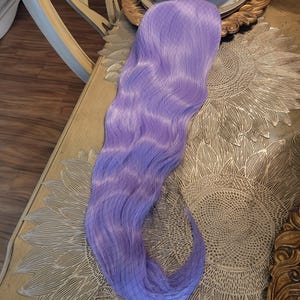 Puede incluir: Una extensi&oacute;n de cabello sint&eacute;tico largo y ondulado en un vibrante color morado lavanda. La pieza de cabello presenta una textura suave y fluida con ondas suaves a lo largo de toda su extensi&oacute;n, mostrando un tono morado claro uniforme.