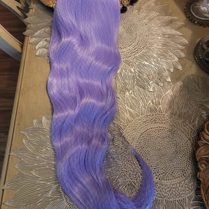 Puede incluir: Una extensi&oacute;n de cabello sint&eacute;tico larga y ondulada en un vibrante color morado lavanda. La pieza de cabello presenta una textura suave y fluida con un patr&oacute;n de ondas ligeras a lo largo de su extensi&oacute;n, terminando en un extremo curvado.