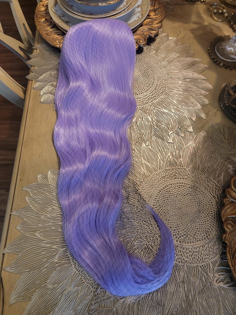 Puede incluir: Una extensi&oacute;n de cabello sint&eacute;tico larga y ondulada en un vibrante color morado lavanda. La pieza de cabello presenta una textura suave y fluida con un patr&oacute;n de ondas ligeras a lo largo de su extensi&oacute;n, terminando en un extremo curvado.