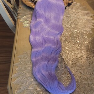 Puede incluir: Una peluca sint&eacute;tica larga y ondulada de un vibrante color morado lavanda. Las fibras capilares son lisas y tienen un ligero brillo, peinadas con ondas suaves y fluidas que se extienden desde la parte superior hasta el extremo curvo.