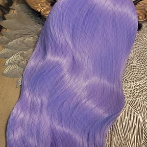 Puede incluir: Una peluca larga y lisa de color lavanda, envuelta en una redecilla negra con patr&oacute;n de rombos. Las fibras capilares sint&eacute;ticas son de un color violeta claro s&oacute;lido y vibrante, con una textura suave y un ligero brillo a lo largo del cabello.