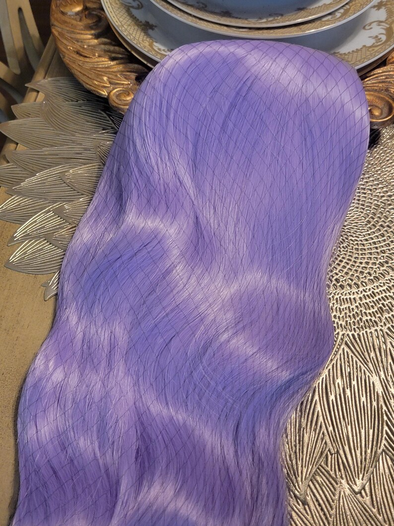 Puede incluir: Una peluca larga y lisa de color lavanda, envuelta en una redecilla negra con patr&oacute;n de rombos. Las fibras capilares sint&eacute;ticas son de un color violeta claro s&oacute;lido y vibrante, con una textura suave y un ligero brillo a lo largo del cabello.