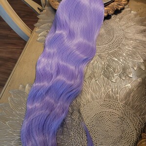 Puede incluir: Una extensi&oacute;n de cabello larga y ondulada en un vibrante color morado lavanda. La pieza de cabello presenta una textura sint&eacute;tica suave con un tono violeta claro constante en toda su longitud, peinada en ondas suaves y fluidas que caen.