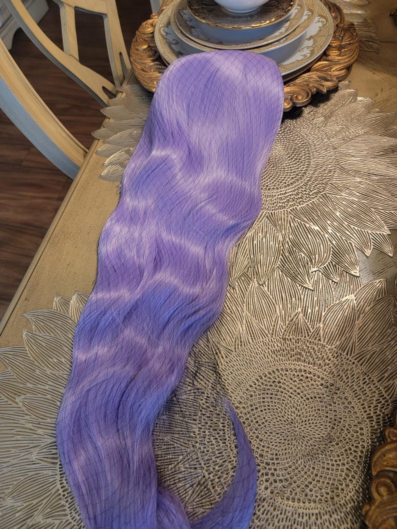 Puede incluir: Una extensi&oacute;n de cabello larga y ondulada en un vibrante color morado lavanda. La pieza de cabello presenta una textura sint&eacute;tica suave con un tono violeta claro constante en toda su longitud, peinada en ondas suaves y fluidas que caen.