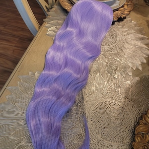 Puede incluir: Una peluca sint&eacute;tica larga y ondulada de un vibrante color morado lavanda. Las fibras capilares presentan una textura suave y fluida con ondas suaves en toda su longitud. Una redecilla fina de color oscuro cubre la parte superior de la peluca.