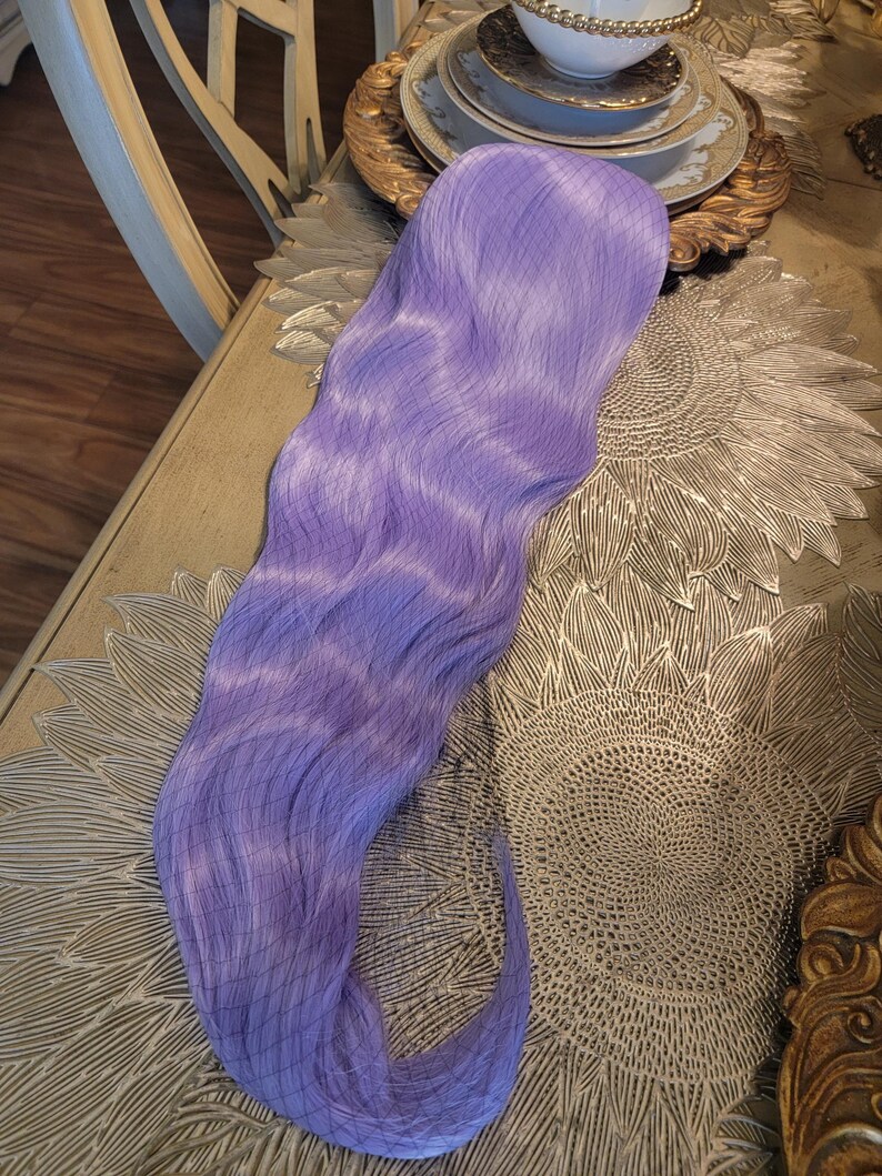 Puede incluir: Una peluca sint&eacute;tica larga y ondulada de un vibrante color morado lavanda. Las fibras capilares presentan una textura suave y fluida con ondas suaves en toda su longitud. Una redecilla fina de color oscuro cubre la parte superior de la peluca.