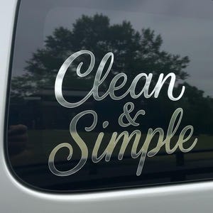 May include: A silver decal with the words "Clean & Simple" in a cursive font is affixed to a car window. The background shows a blurred view of trees and a cloudy sky.