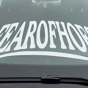 May include: White vinyl decal with the text "FEAROFHOES" in a stylized font, applied to a car windshield. The decal is curved to fit the windshield's shape, and the text is centered.