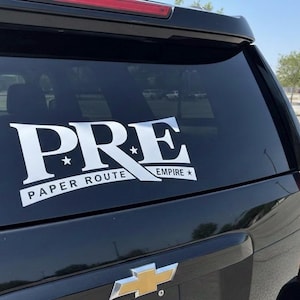May include: A black SUV with a white decal on the rear window. The decal reads "PRE PAPER ROUTE EMPIRE" in a bold, stylized font. The vehicle's taillight is visible on the right side, and a gold Chevrolet emblem is at the bottom.