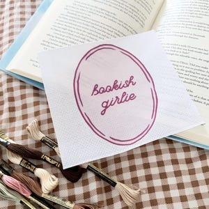 Op de afbeelding: Een witte kruissteek met de woorden "bookish girlie" in een paarse ovaal, rustend op een open boek. Verschillende spoelen borduurgaren in neutrale kleuren zijn eronder gerangschikt, op een bruin-wit geblokt oppervlak.