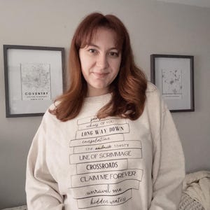 Puede incluir: Una persona lleva una sudadera crema con texto negro. El texto incluye frases como "Long Way Down" y "Claim Me Forever". Dos mapas enmarcados son visibles en el fondo. La persona tiene el pelo castaño rojizo.