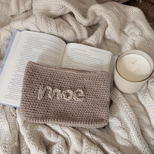 May include: A knitted headband in a light brown hue, with the name "moe" stitched in a lighter color. The headband rests on an open book, next to a lit candle, all on a textured, cream-colored blanket.