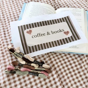 May include: An open book with a white canvas featuring the text "coffee & books" and two heart illustrations. Several skeins of embroidery floss in shades of brown, pink, and green are arranged on a brown and white checkered surface.